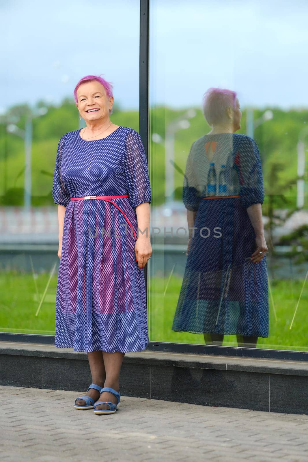 Old woman with cheerful smile in blue dress standing near the modern building with glass walls