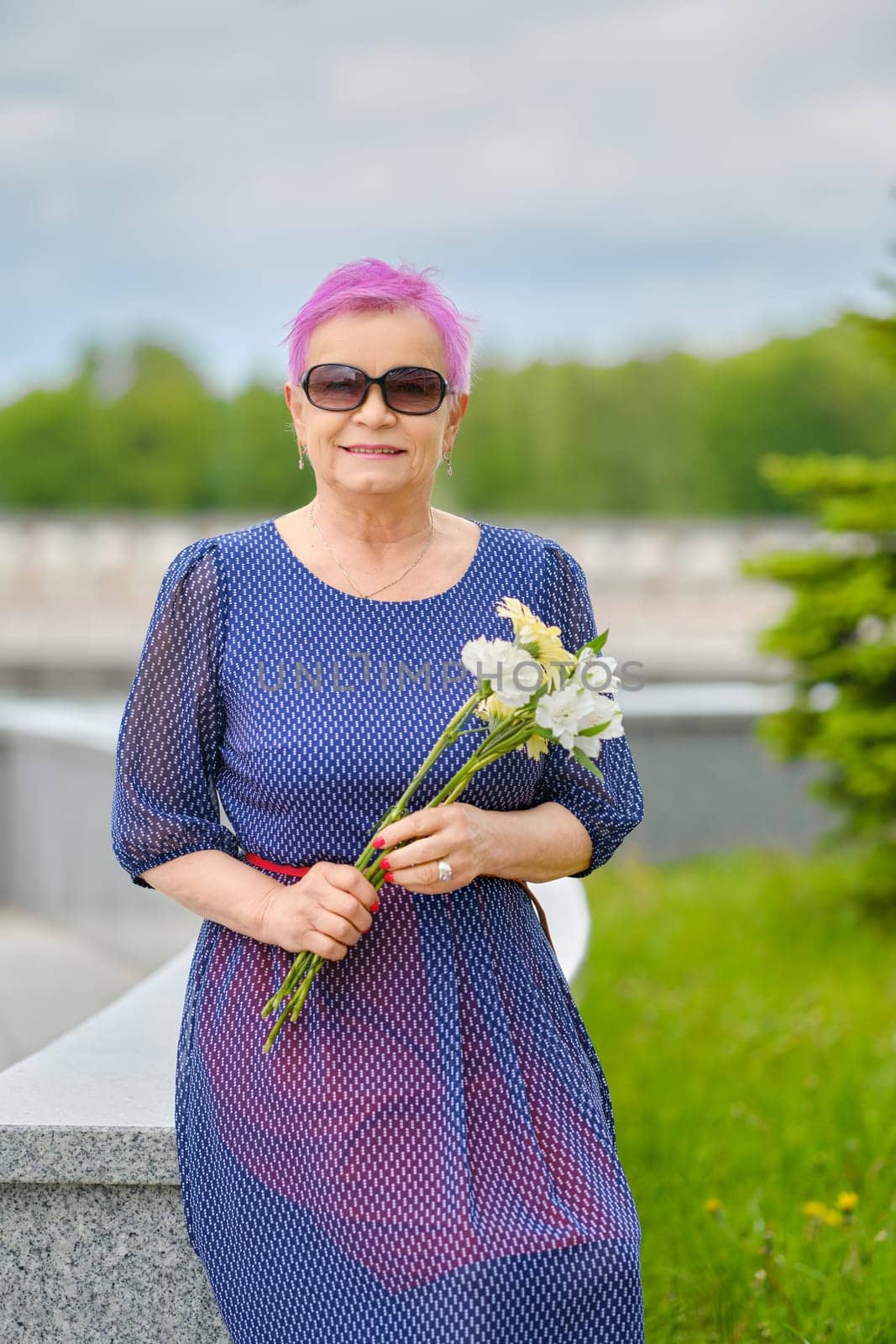 Portrait of smiling mature woman with short pink hair standing near fir-tree on the street