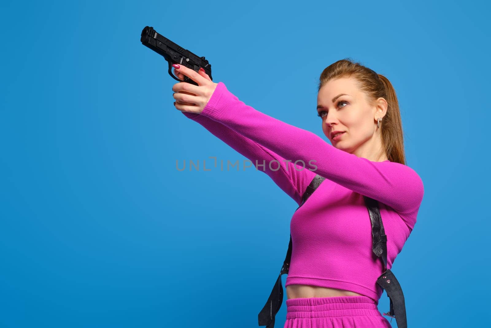 Stylish woman in purple knitted pantsuit aims a gun over blue studio background. Fashion police and accurate hit on target concept.