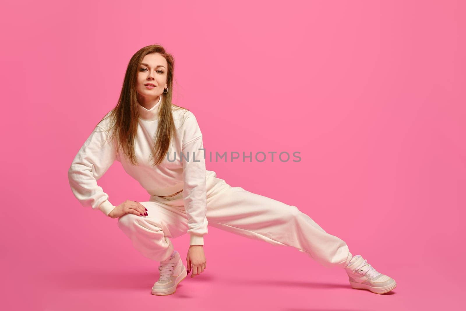 Active woman in white sportswear sits on squats and leaning on one leg over pink studio background