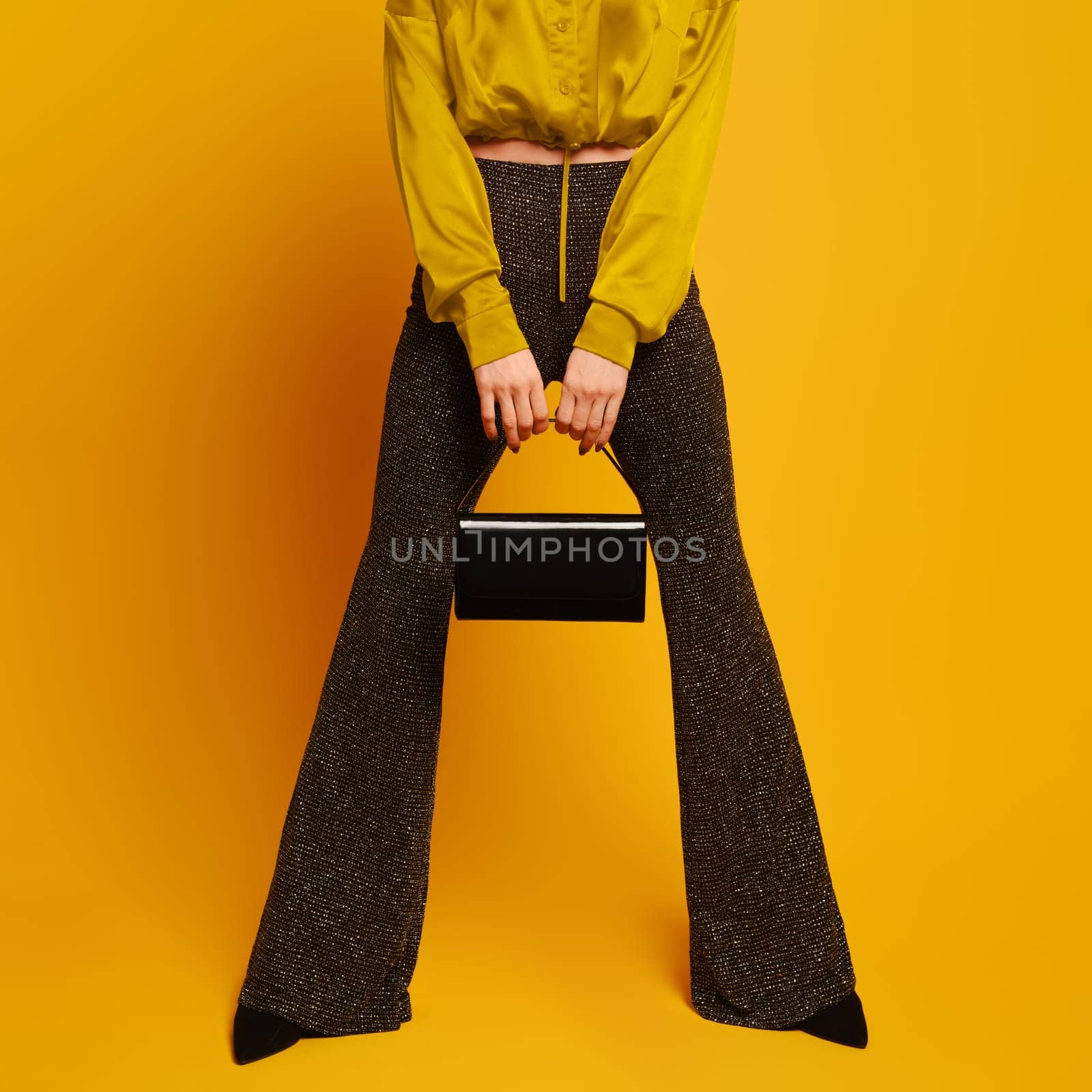 Female legs in flared trousers stand wide apart and hands hold a purse between them on yellow studio background