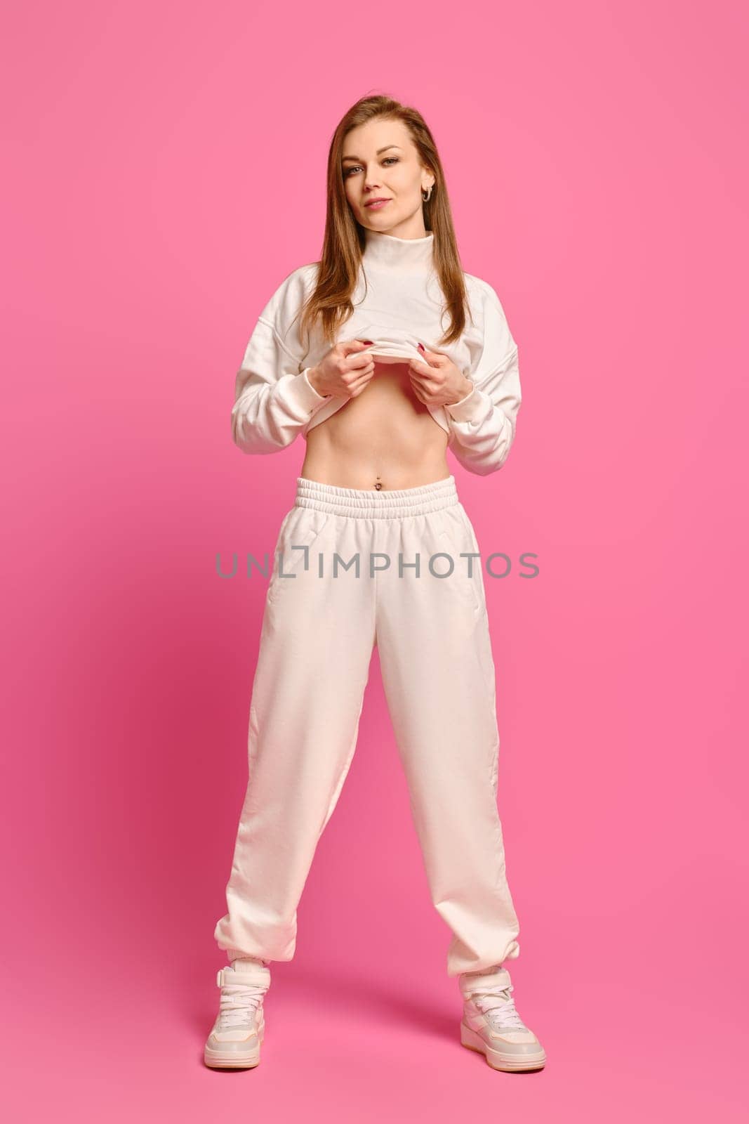 Beautiful woman lifted her sweatshirt and shows a flat tummy standing over bright pink background