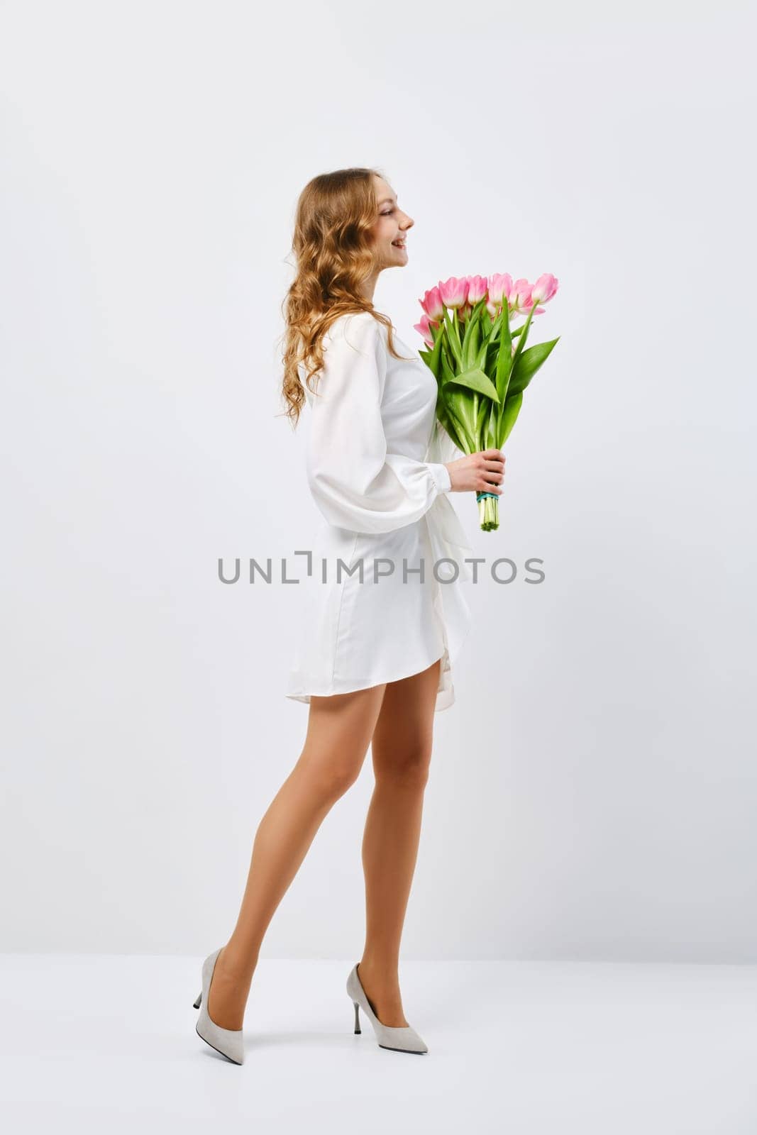 Young happy woman in white dress turning to the side with bouquet of tulips over white background