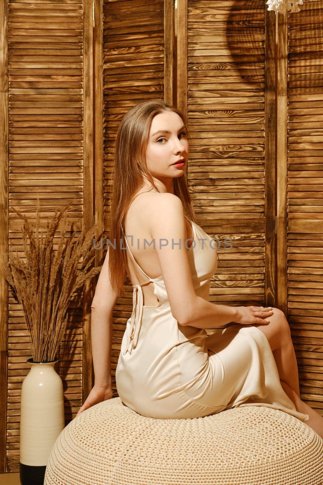Beautiful woman in silky spaghetti strap dress sitting on padded stool in profile