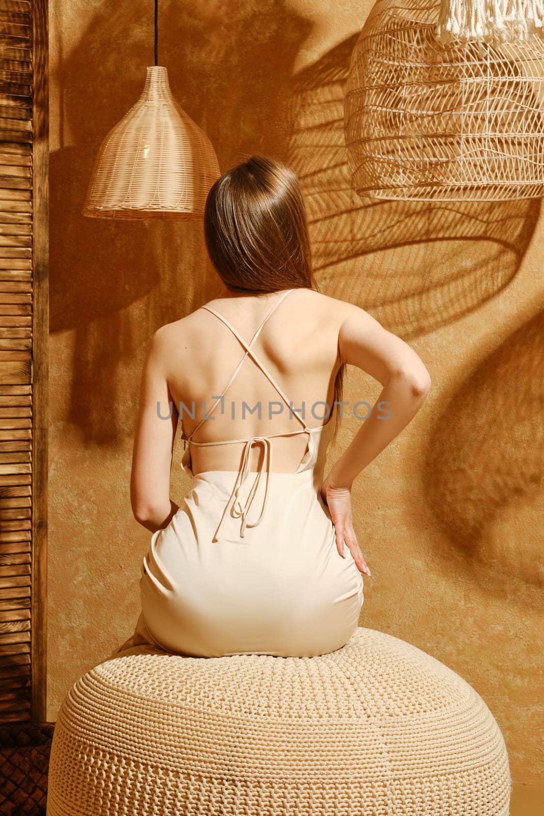Rear view of young woman in silky spaghetti strap dress sitting on padded stool