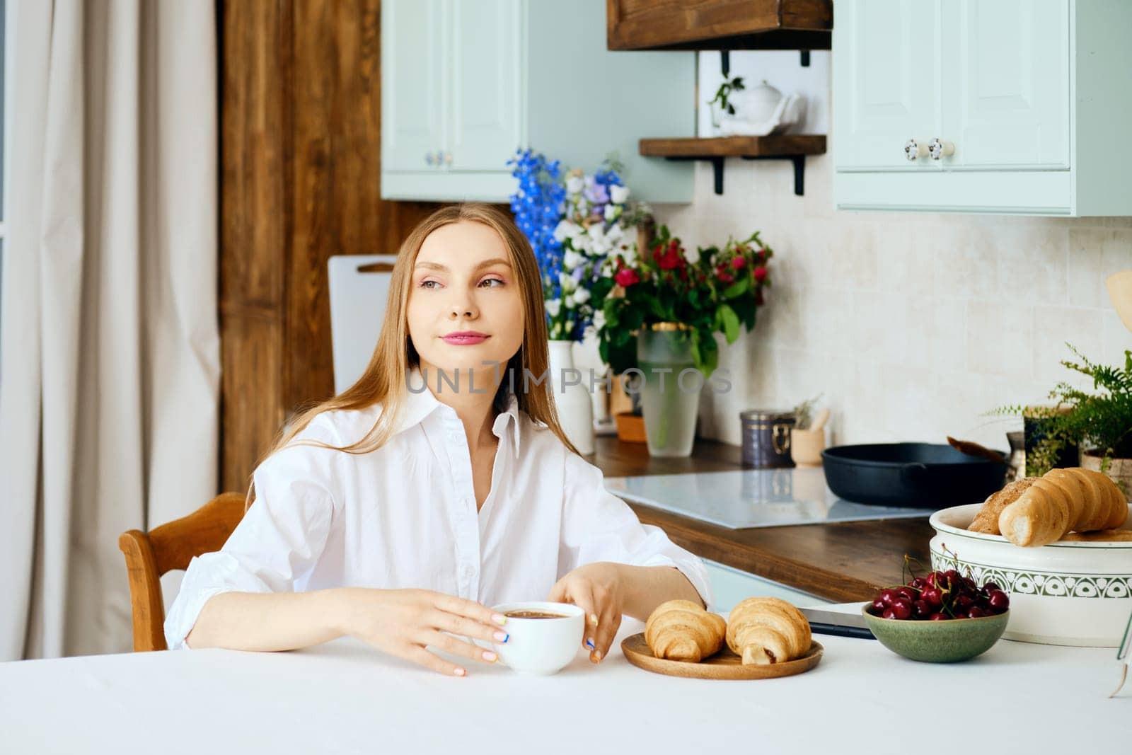 Positive young woman sits behind the kitchen table with cup of coffee