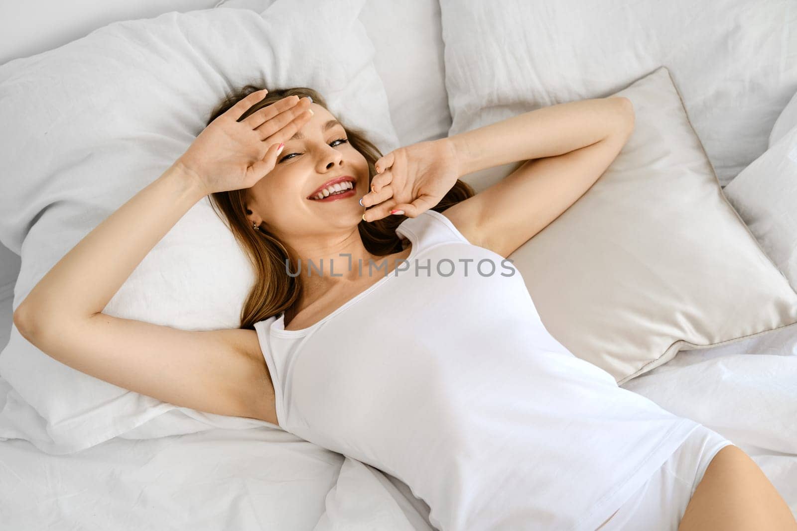 Young positive woman in white tank top and panties in bed covers face with hands and smiling