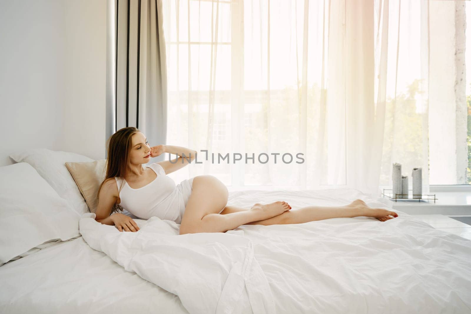 Young positive woman in white tank top and panties in bed by the window