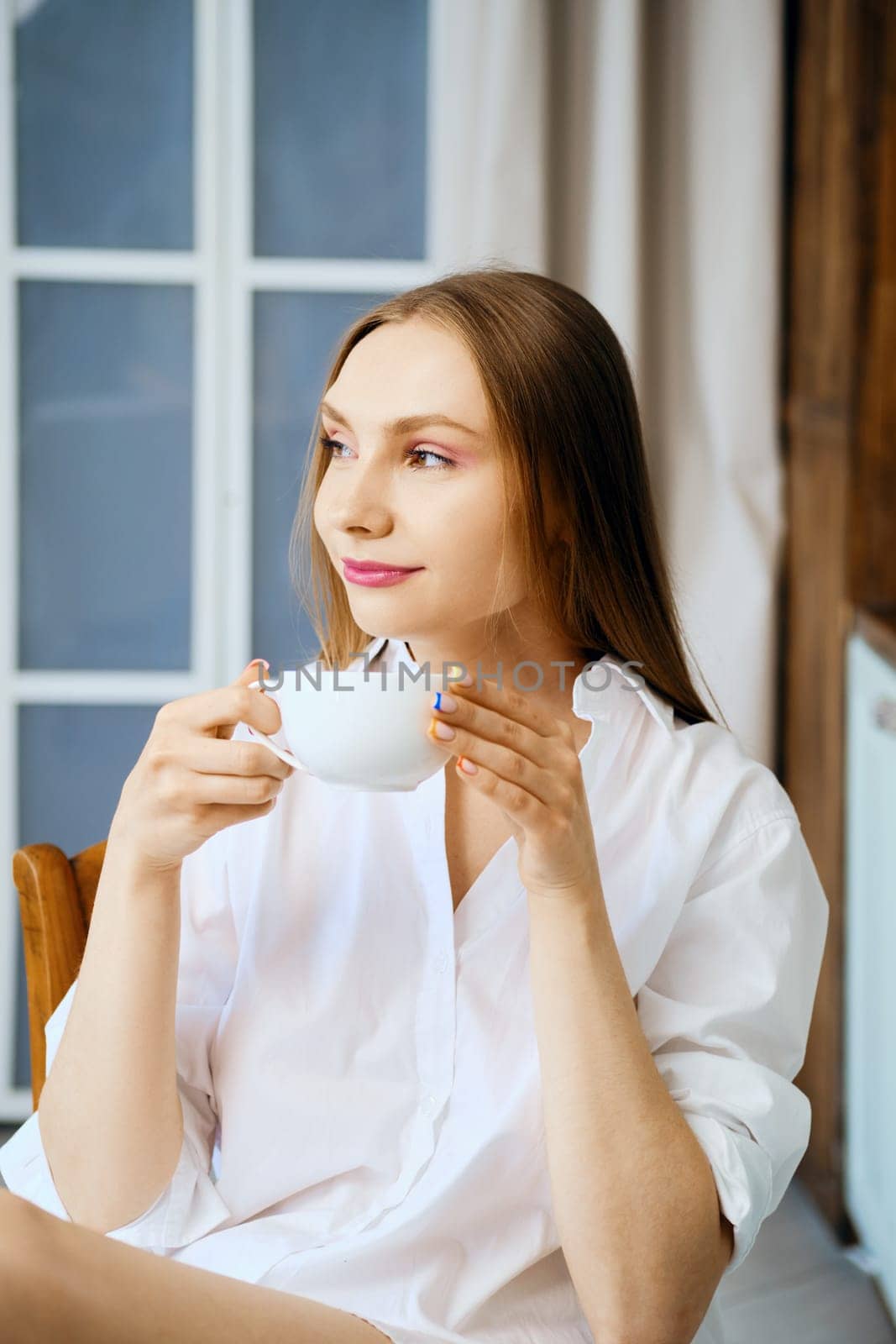 Pensive young woman in white shirt in the kitchen holds cup of coffee