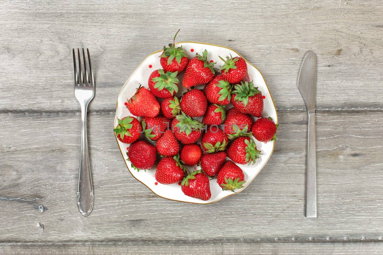 Tabletop view, plate with strawberries on gray wood boards desk, fork and knife next to it - ready to eat healthy snack concept.