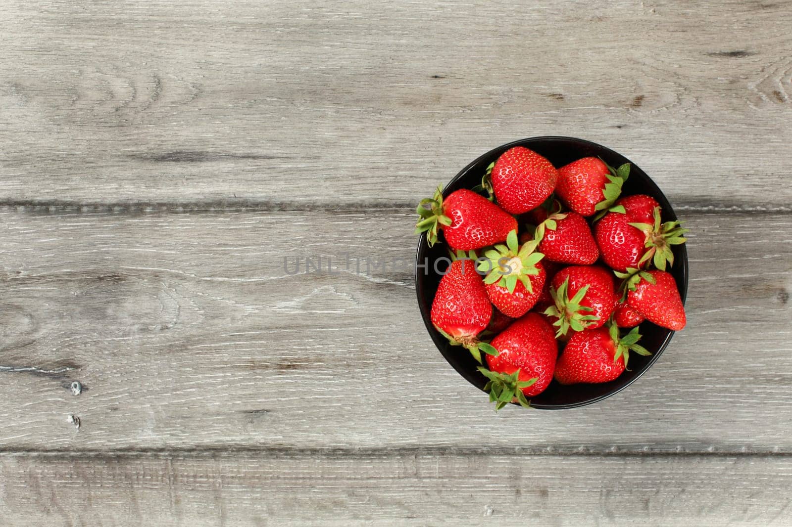 Tabletop view, small black ceramic bowl with strawberries, gray wood desk under. Space for text on left side.