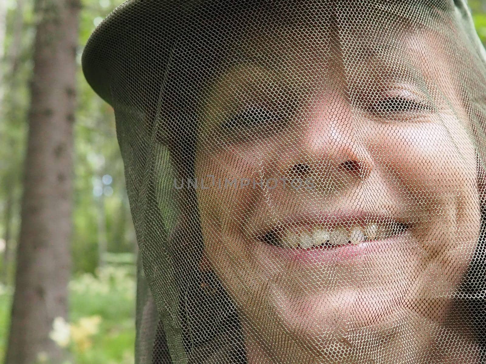 A woman in her 40s wearing a mosquito headdress smiles and looks at the camera. Cap and mesh against insects, mosquitoes, bees, wasps. Hunting and fishing in the spruce forest.