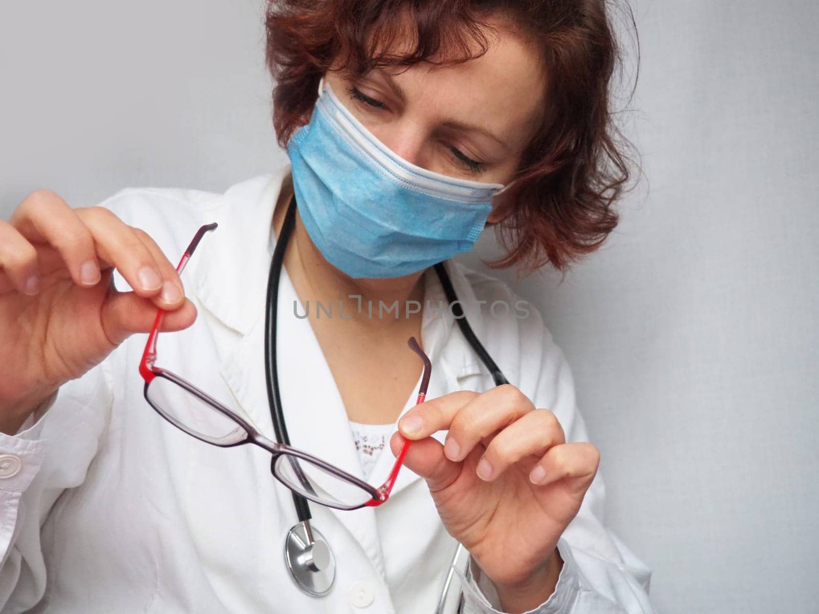 Doctor with stethoscope examining red-rimmed glasses. Medicine and healthcare. A frowning, concentrated face in a protective mask. An ophthalmologist or cardiologist. Gray background.