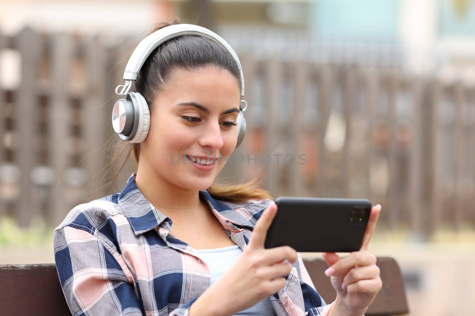 Happy teen wearing headphones watching media on phone in a bench