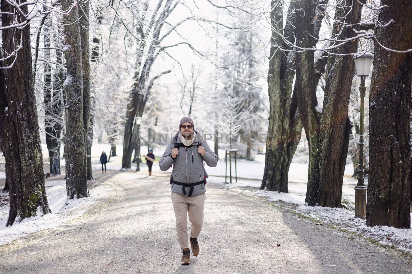 Sporty father carrying his infant son wearing winter jumpsuit and cap in backpack carrier walking in city park in winter.