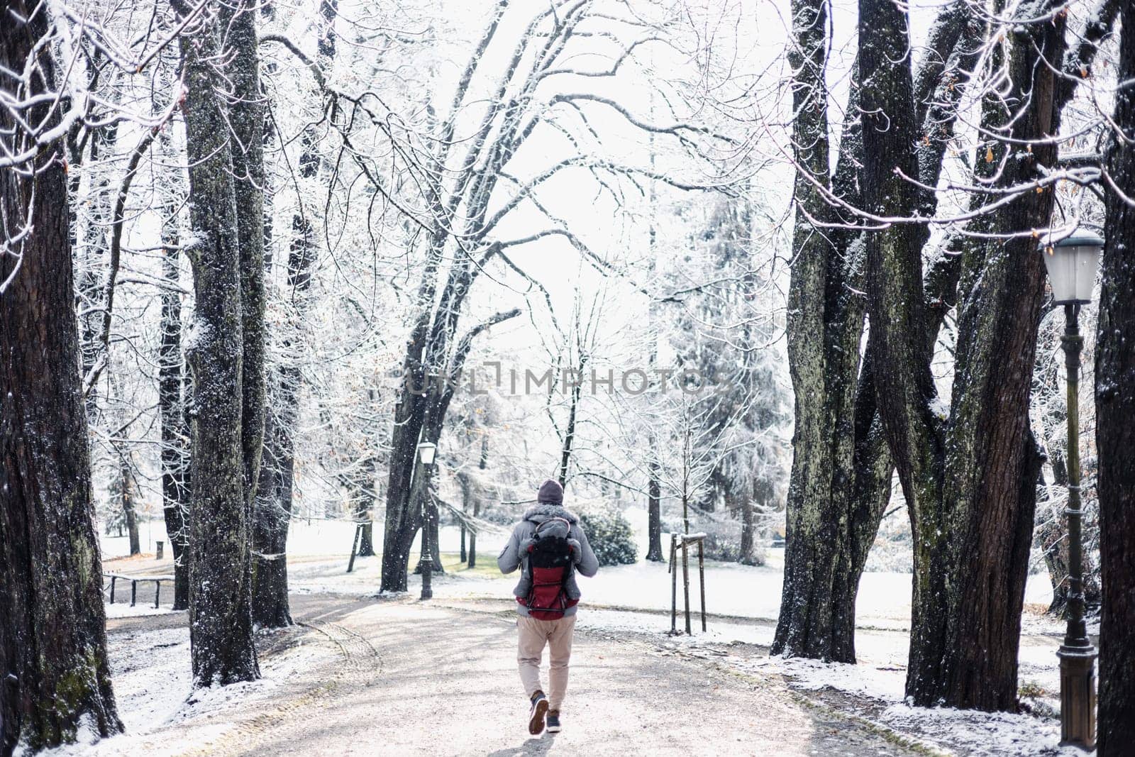 Rear view of sporty father carrying his infant son wearing winter jumpsuit and cap in backpack carrier walking in city park in winter.