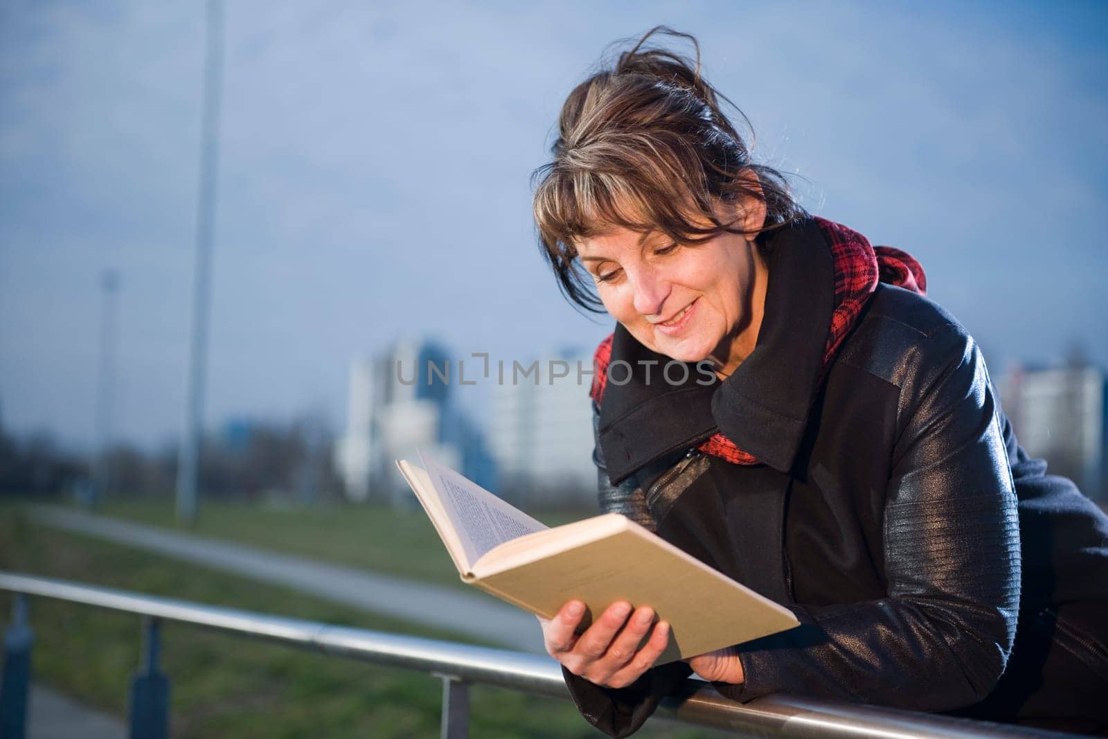Side-body view of a mature woman with black leather jacket leaning on a railing in a book reading against a fuzzy urban background
