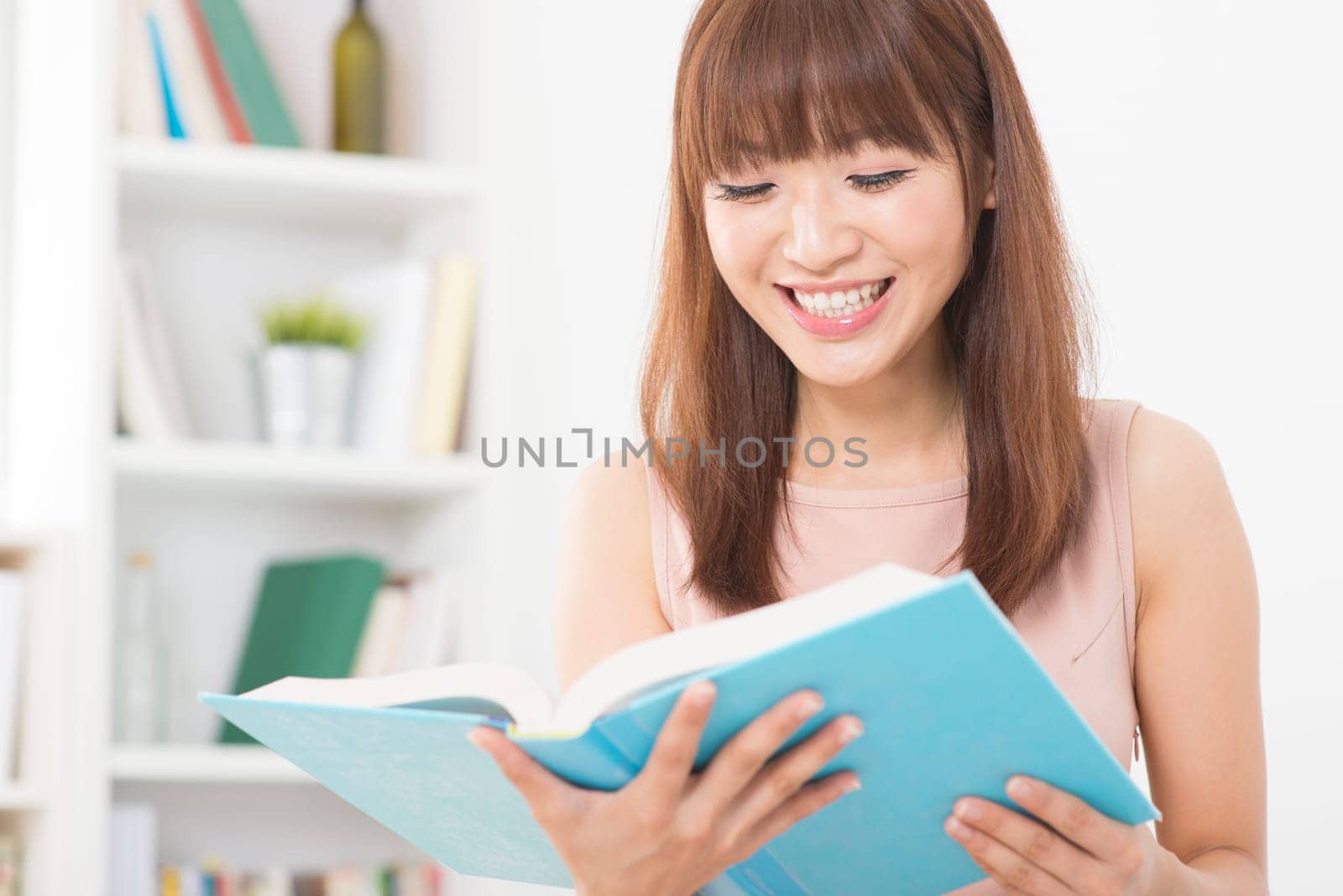 Portrait of happy Asian college girl laughing while reading on book. Young woman at school library.