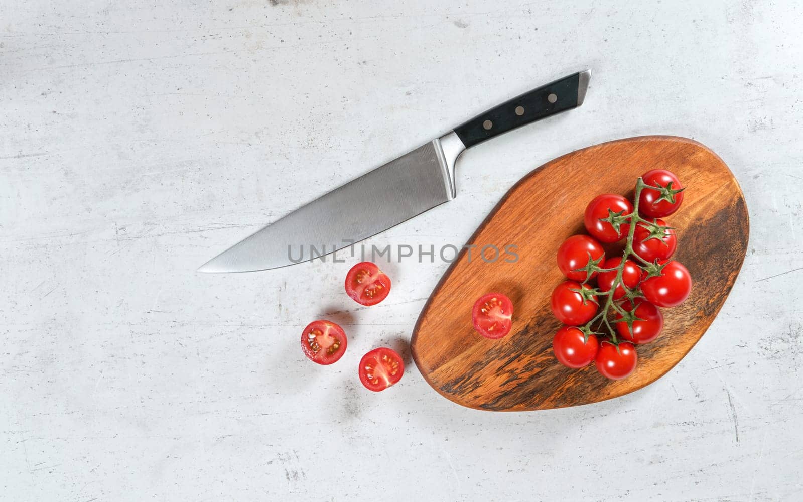Vibrant small red tomatoes with green vines on wooden chopping board, large chef knife near, white stone table under, view from above.