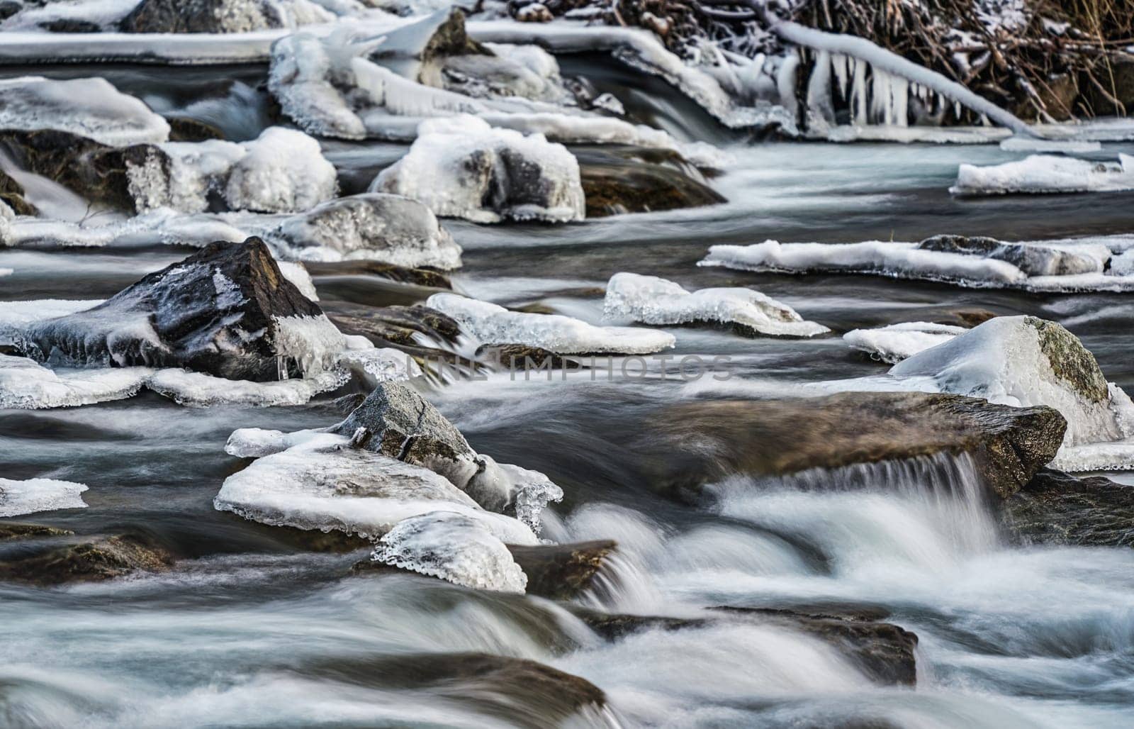 Winter river flowing over snow and ice covered stones, closeup detail, long exposure photo.