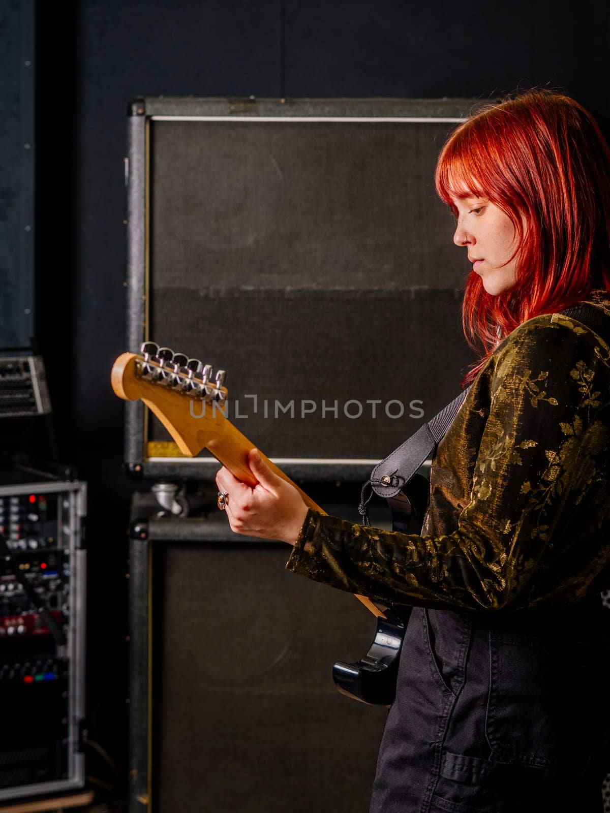 Young redhead woman playing electric guitar in front of large speaker cabinets.