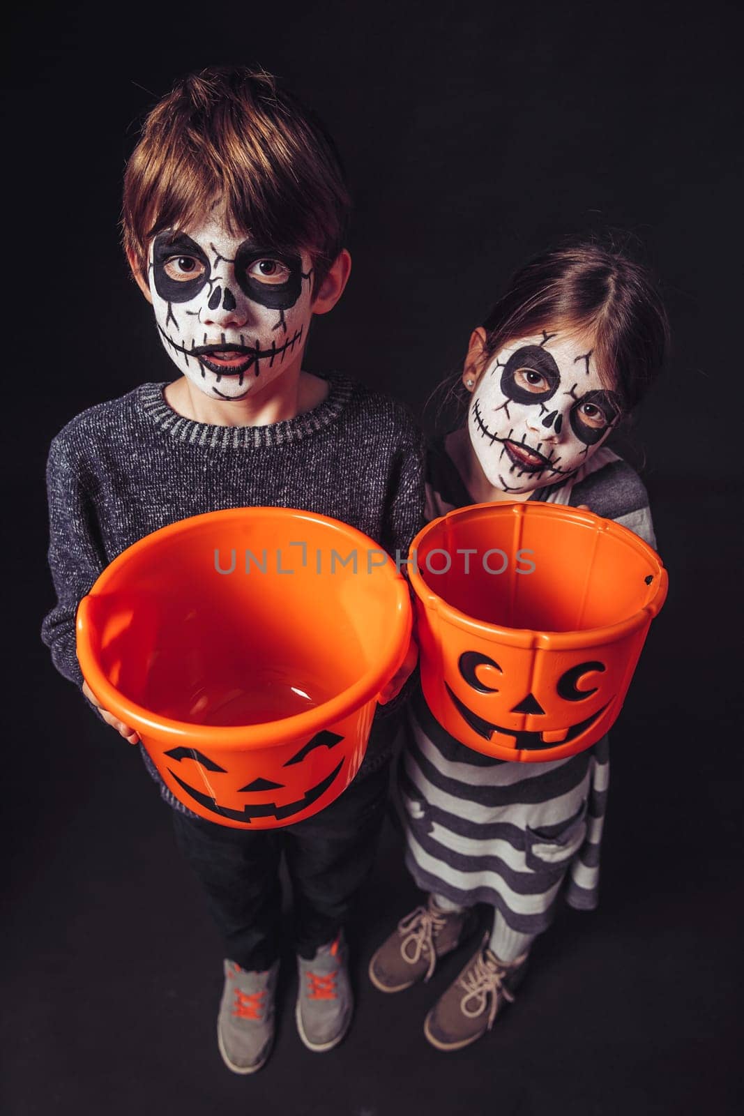 Two children with skull makeup holding pumpkin pails at Halloween.