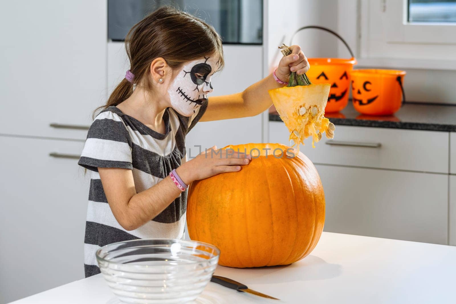 Young girl with skull face paint cutting a pumpkin in their kitchen.