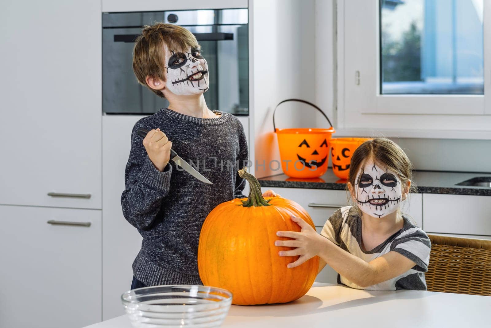 Two young children with skull face paint cutting a pumpkin in their kitchen.