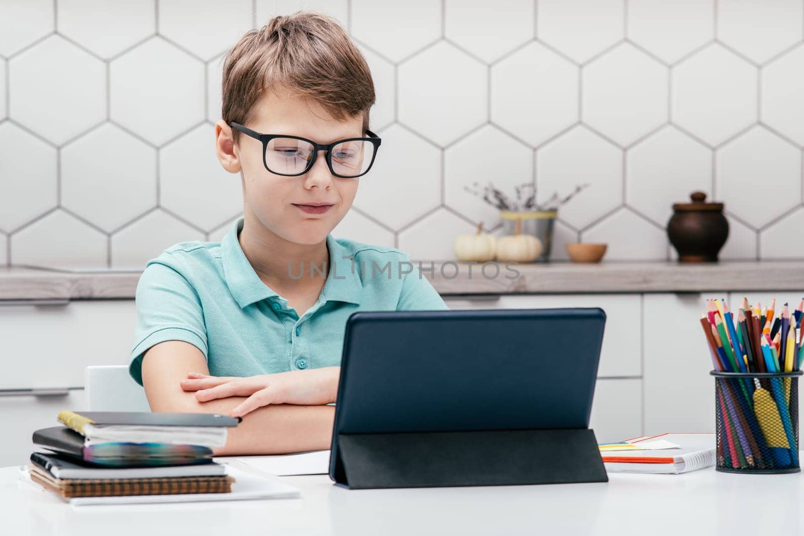 Portrait of young preteen calm boy wearing glasses, sitting with folded hands at desk near notepads, color pencils, looking at screen of tablet, having online lesson. Technology, remote education.