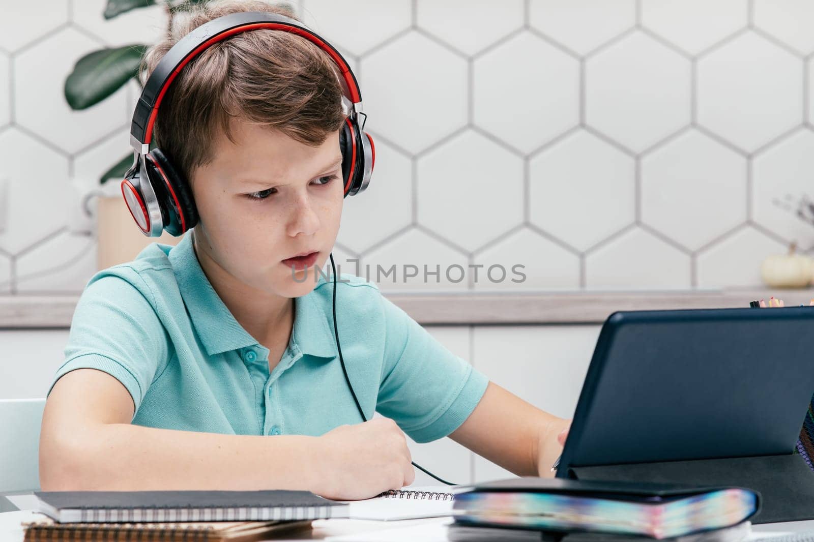 Portrait of young preteen frowning boy wearing T-shirt, headphones, sitting at desk near notebooks, looking at screen of tablet, having online lesson, listening. Remote education, distance learning.