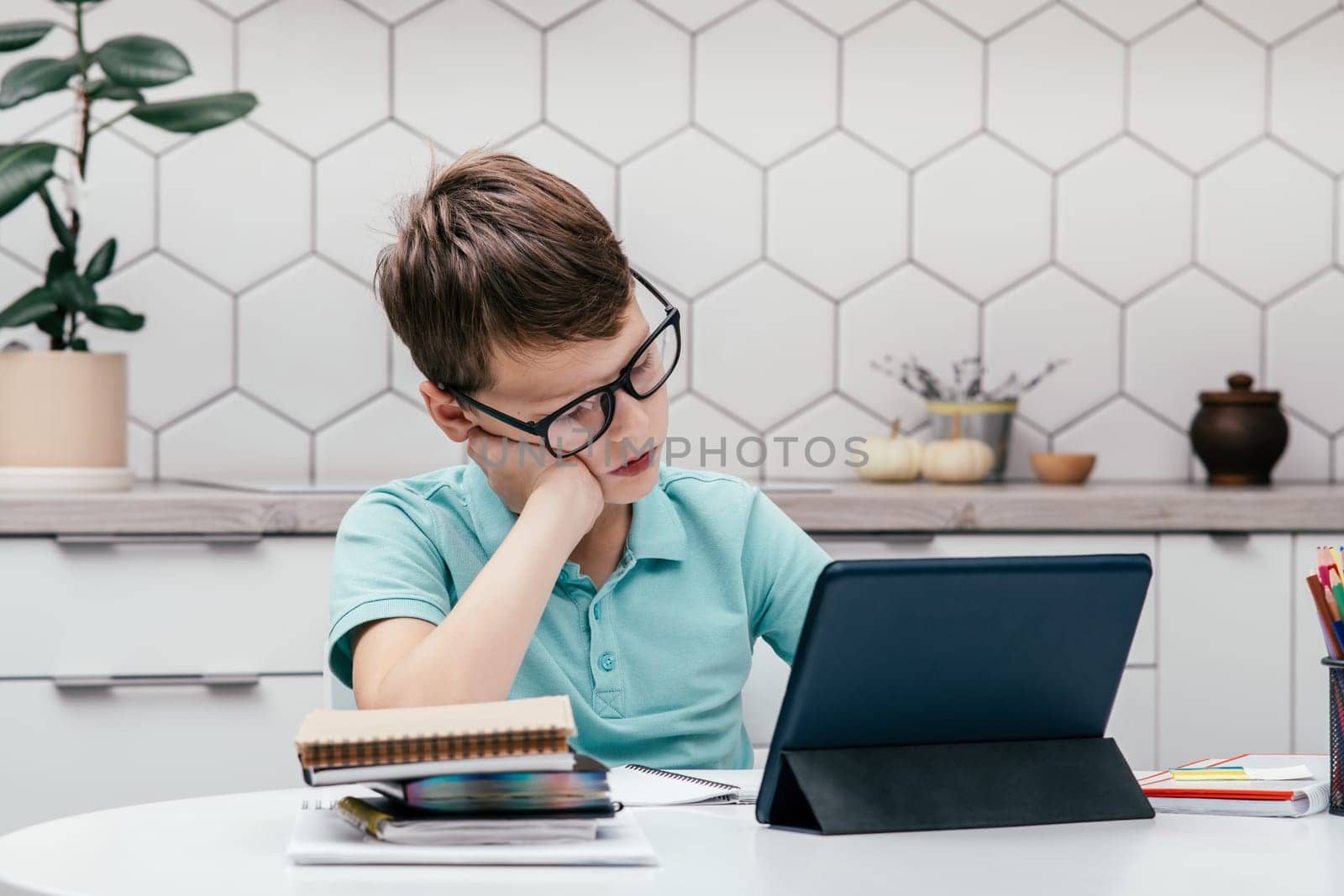 Portrait of young preteen concentrated boy wearing blue T-shirt, glasses, sitting at desk near notepads, reading book near tablet, having online lesson. E-learning, technology, remote education.