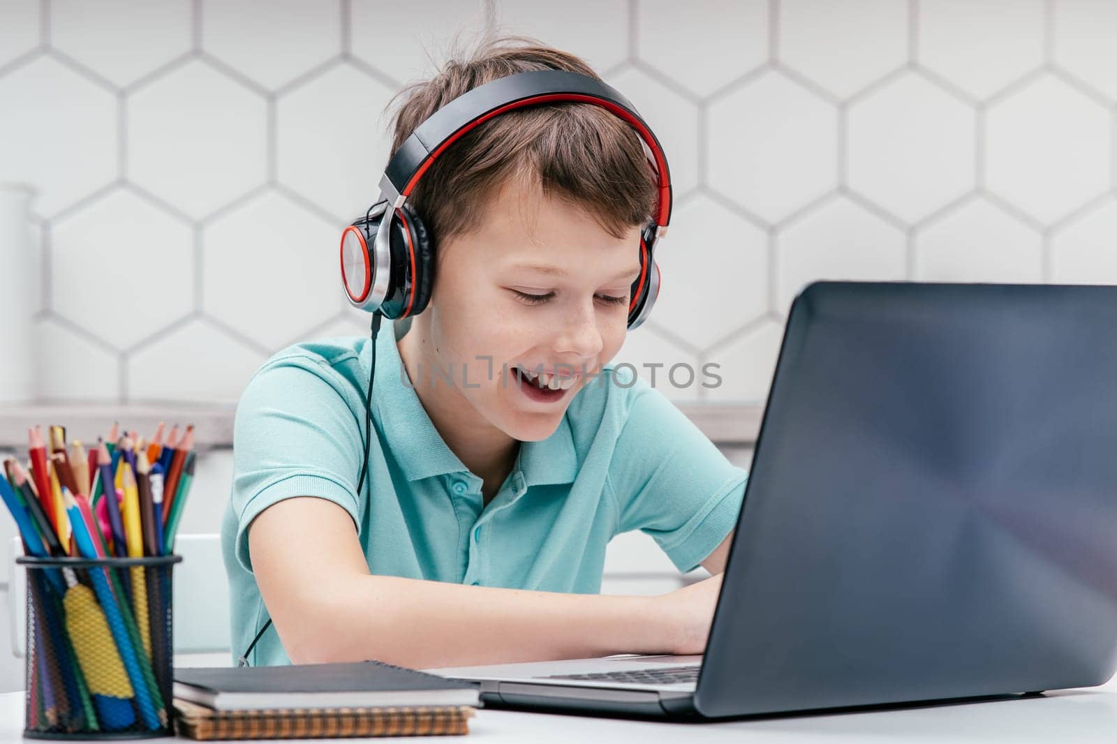 Portrait of young preteen happy boy wearing T-shirt, headphones, sitting at desk in front of laptop, typing, communicating, playing games, having fun. Gaming, technology, e-learning, social media.