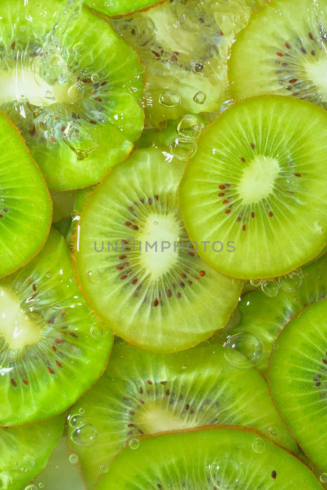 Close-up view of the green kiwi fruit slices in water background. Texture of cooling fruit drink with macro bubbles on the glass wall. Flat design. Vertical image.