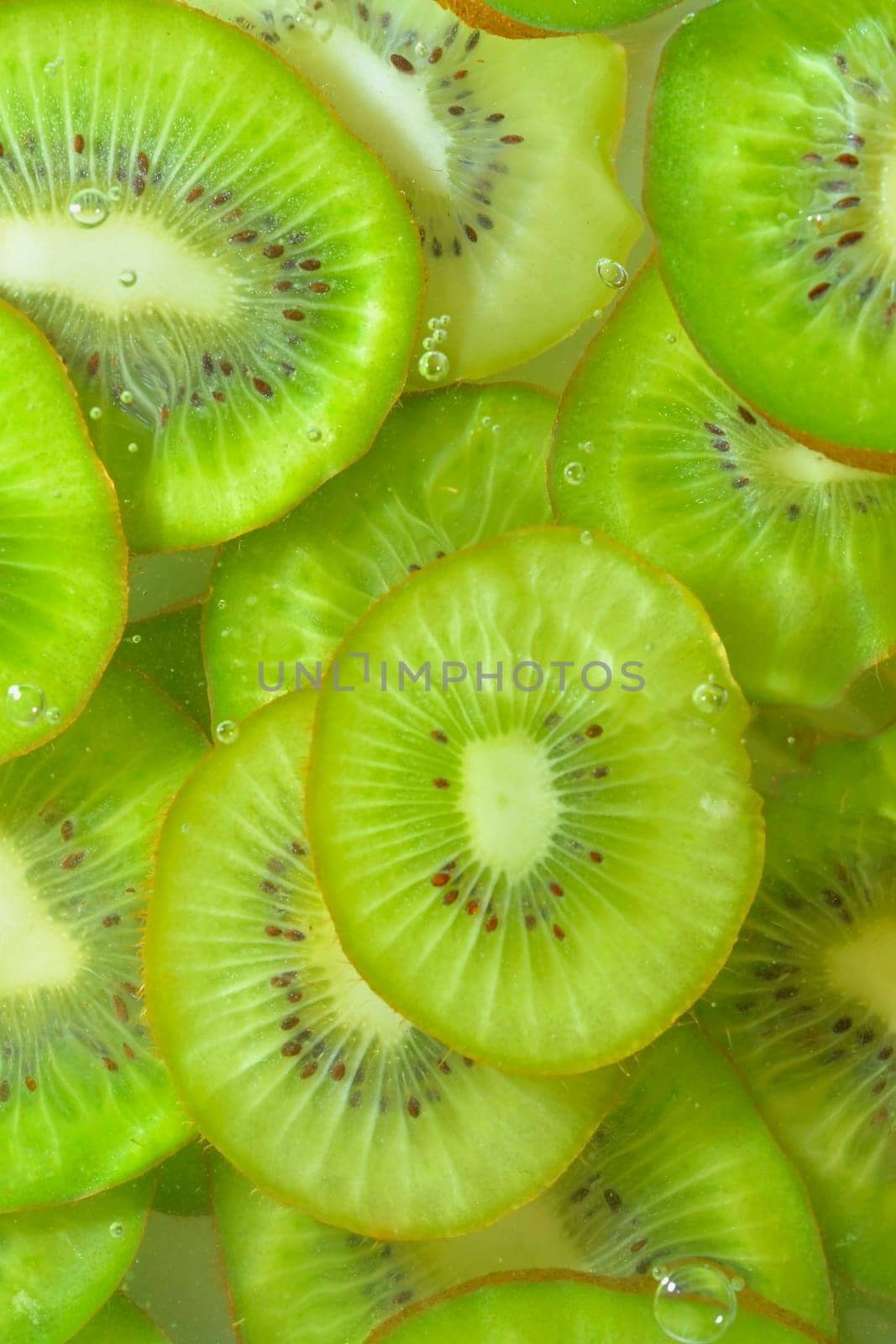 Close-up view of the green kiwi fruit slices in water background. Texture of cooling fruit drink with macro bubbles on the glass wall. Flat design. Vertical image.