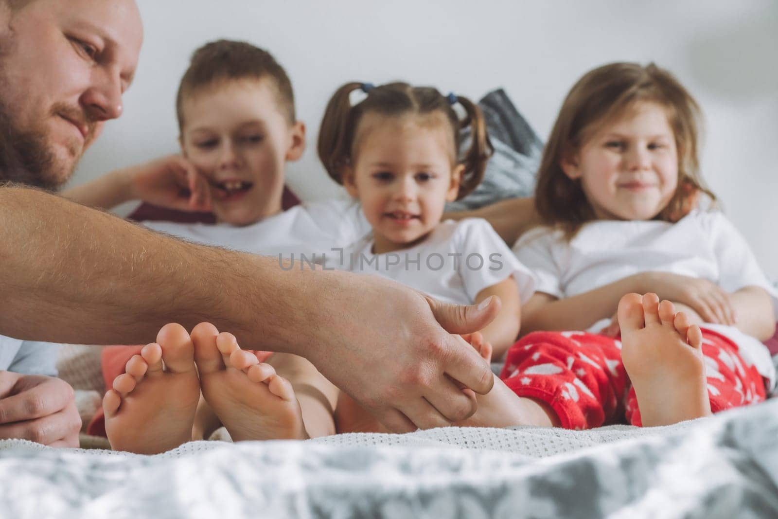 Father plays with three children on bed. Dad tickles kids feet. Family of daddy, two girls and boy