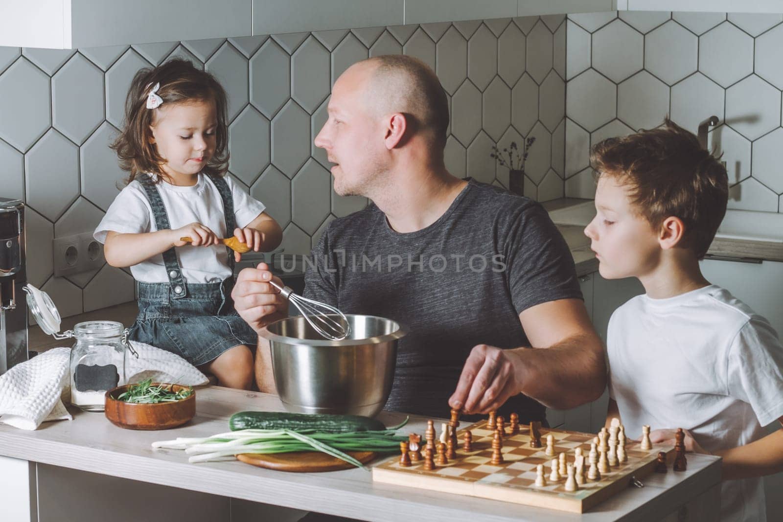 Father whips an omelet with a whisk, playing chess with son and talking to daughter. Man doing household chores.