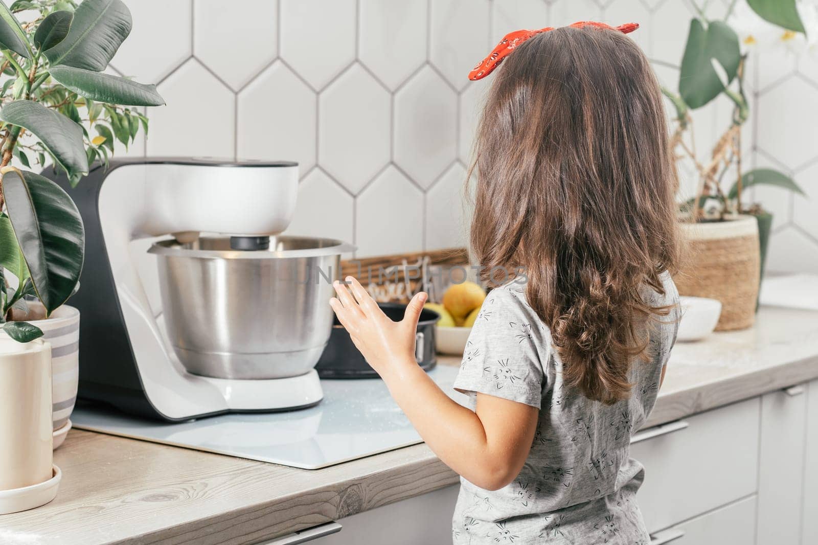 Little dark-haired girl 3 years old in red headband bakes apple pie in kitchen. Child stands near planetary mixer. Children help on household chores. Kid cooking food. Back view
