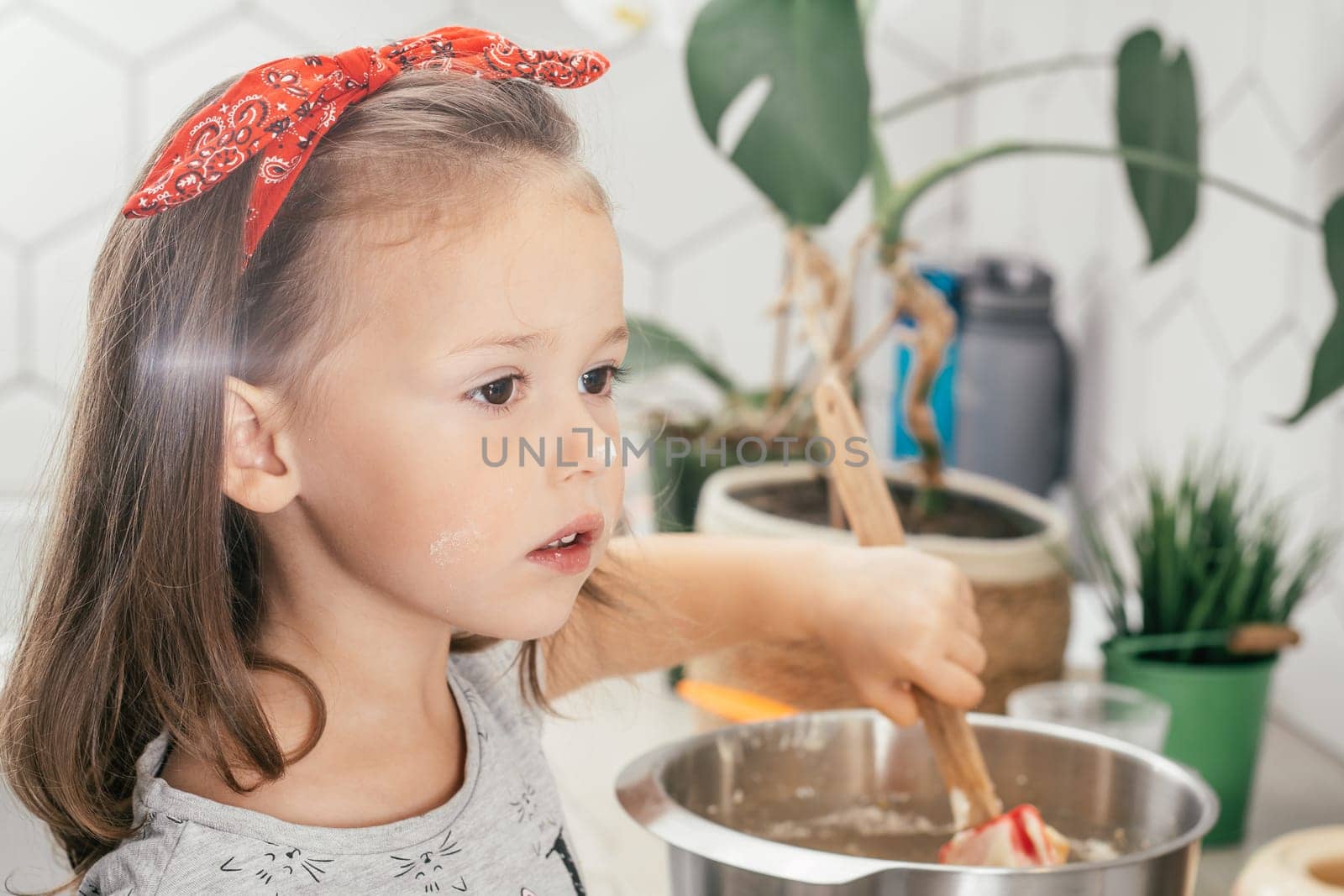 Little dark-haired girl 3 years old in red headband bakes apple pie in kitchen. Child mixes dough in bowl. Children help on household chores. Kid cooking food