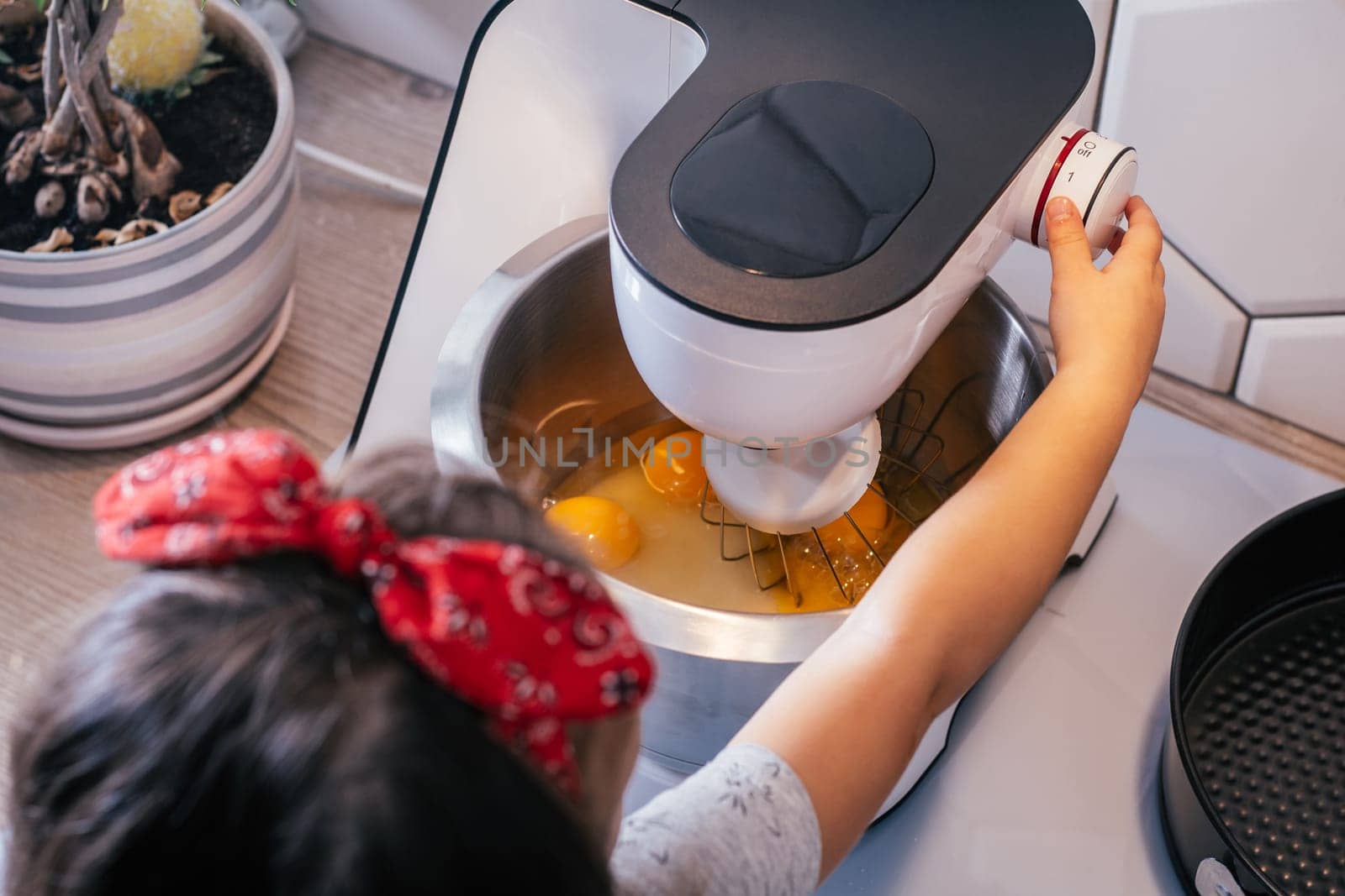 Little dark-haired girl 3 years old in red headband bakes apple pie in kitchen. Child turns on planetary mixer. Children help on household chores. Kid cooking food. Top view