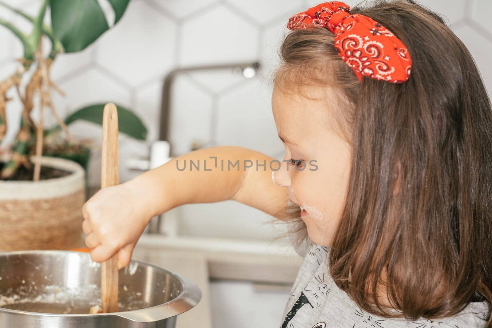Little dark-haired girl 3 years old in red headband bakes apple pie in kitchen. Child mixes dough in bowl. Children help on household chores. Kid cooking food