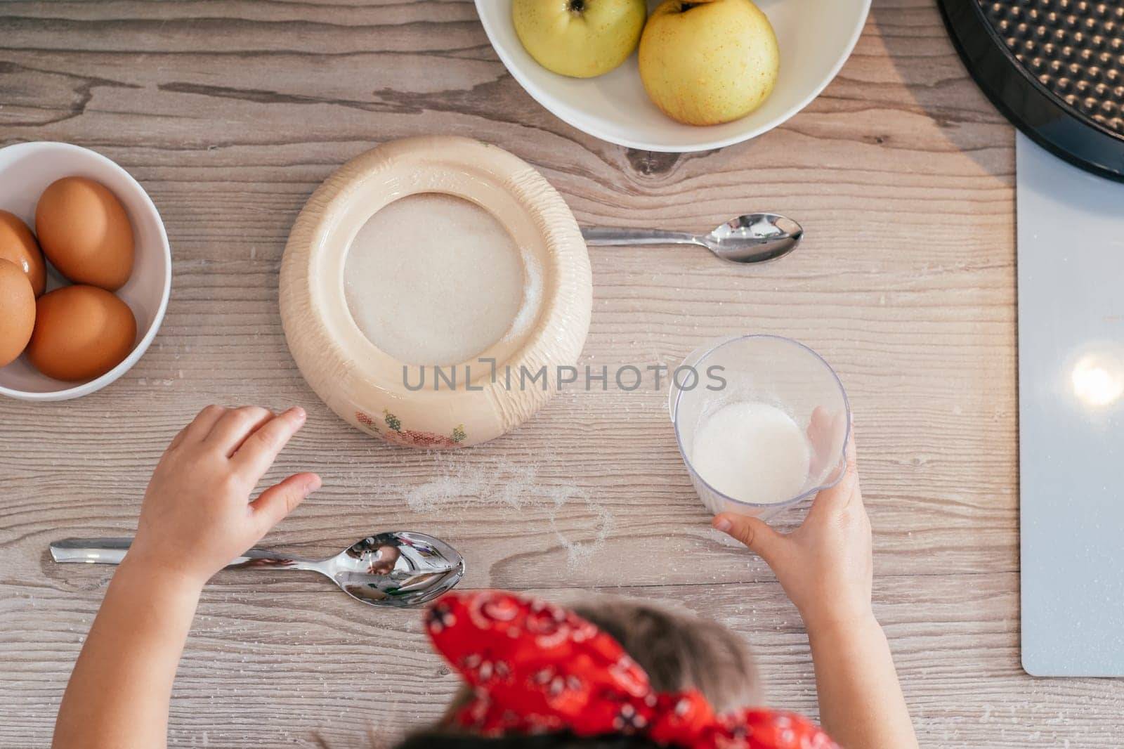 Hands of little girl in red headband bake apple pie in kitchen. Child puts sugar in measuring cup. Children help on household chores. Kid cooking food. Top view