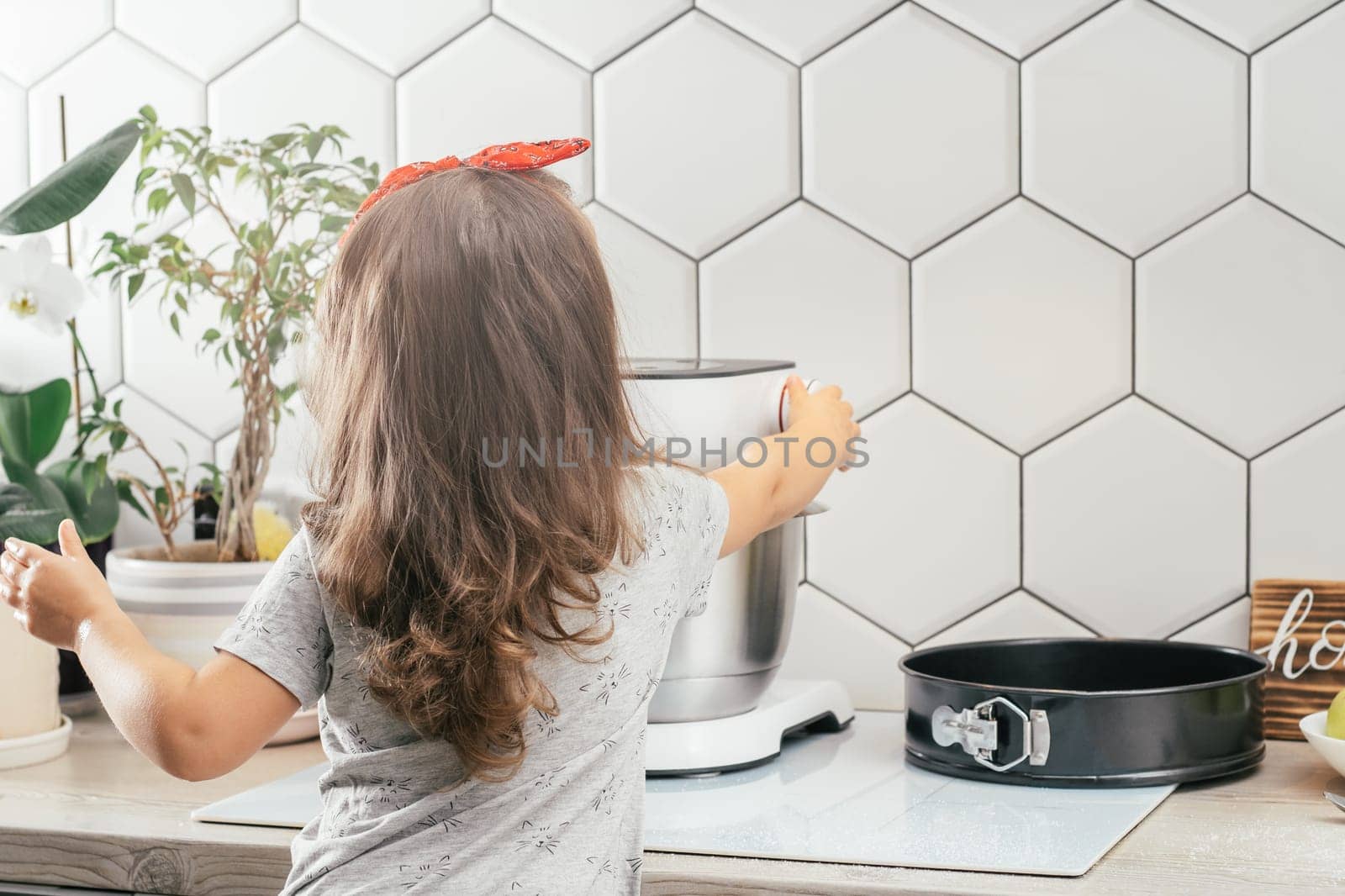 Little dark-haired girl 3 years old in red headband bakes apple pie in kitchen. Child turns on planetary mixer. Children help on household chores. Kid cooking food. Back view