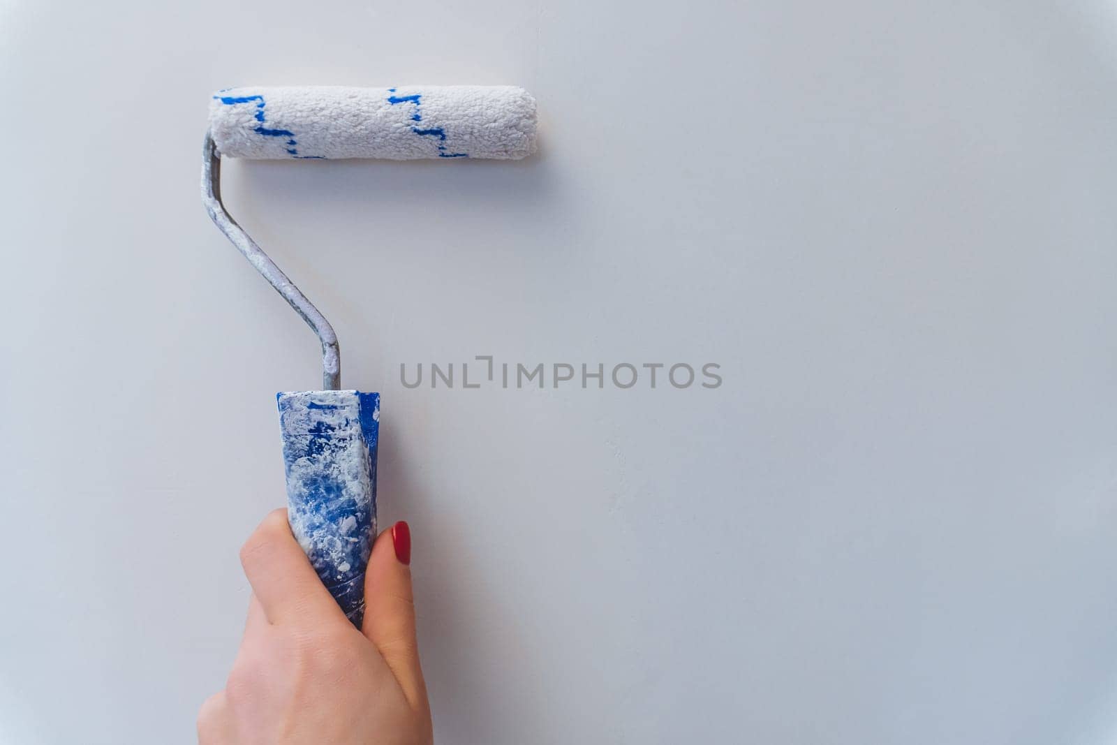 Female hand with red manicure holds roller and paints wall white. Home renovation and improvement.