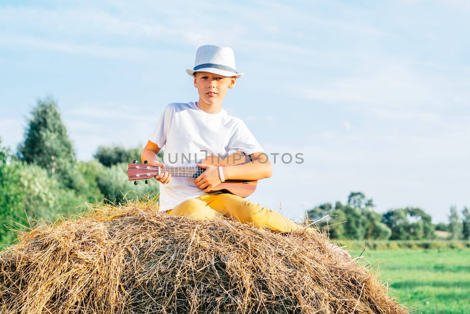 Portrait of barefoot boy in hat on haystack in field. Playing small guitar, ukulele. Light sunny day. Cheerful and music concept. Outdoor activity. Trees on background, Countryside