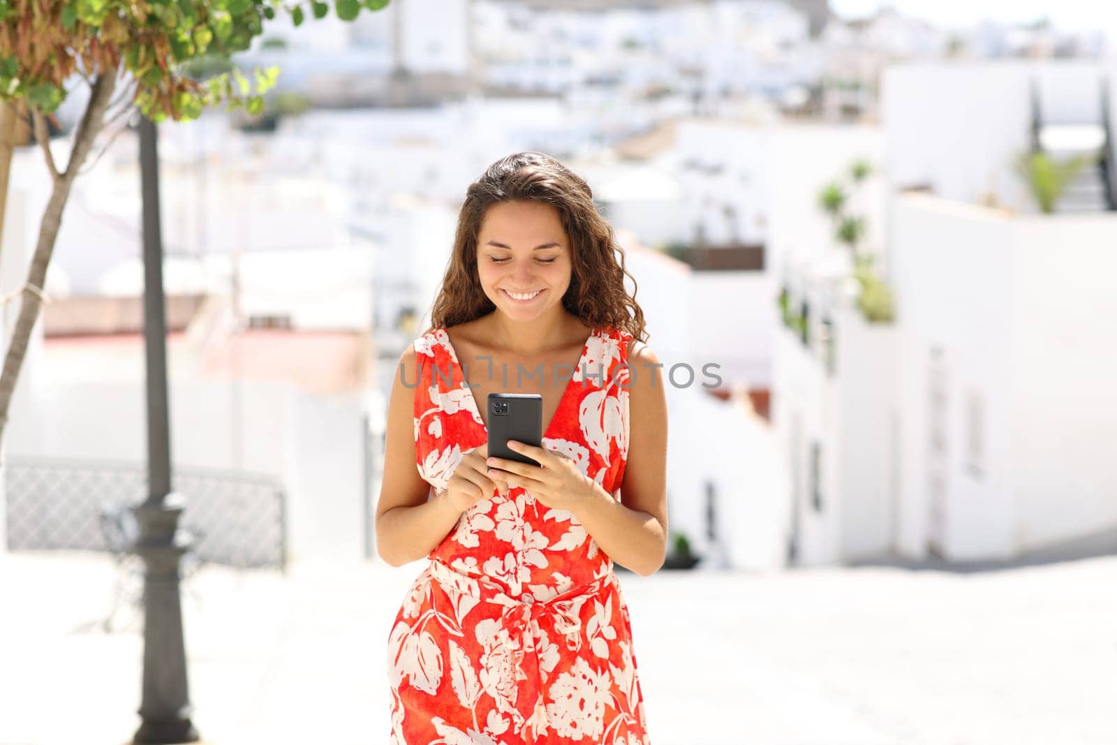 Front view portrait of a happy tourist using smart phone walking in a white spanish town