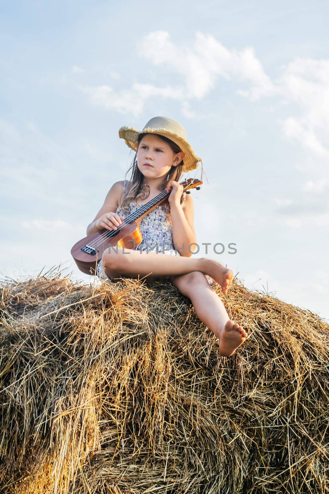Nice little girl sit at top of haystack dangling legs and play ukulele. Carefree child resting on hayrick, from below view. Outdoor walking. Beautiful sky. Summer vacation, countryside lifestyle.