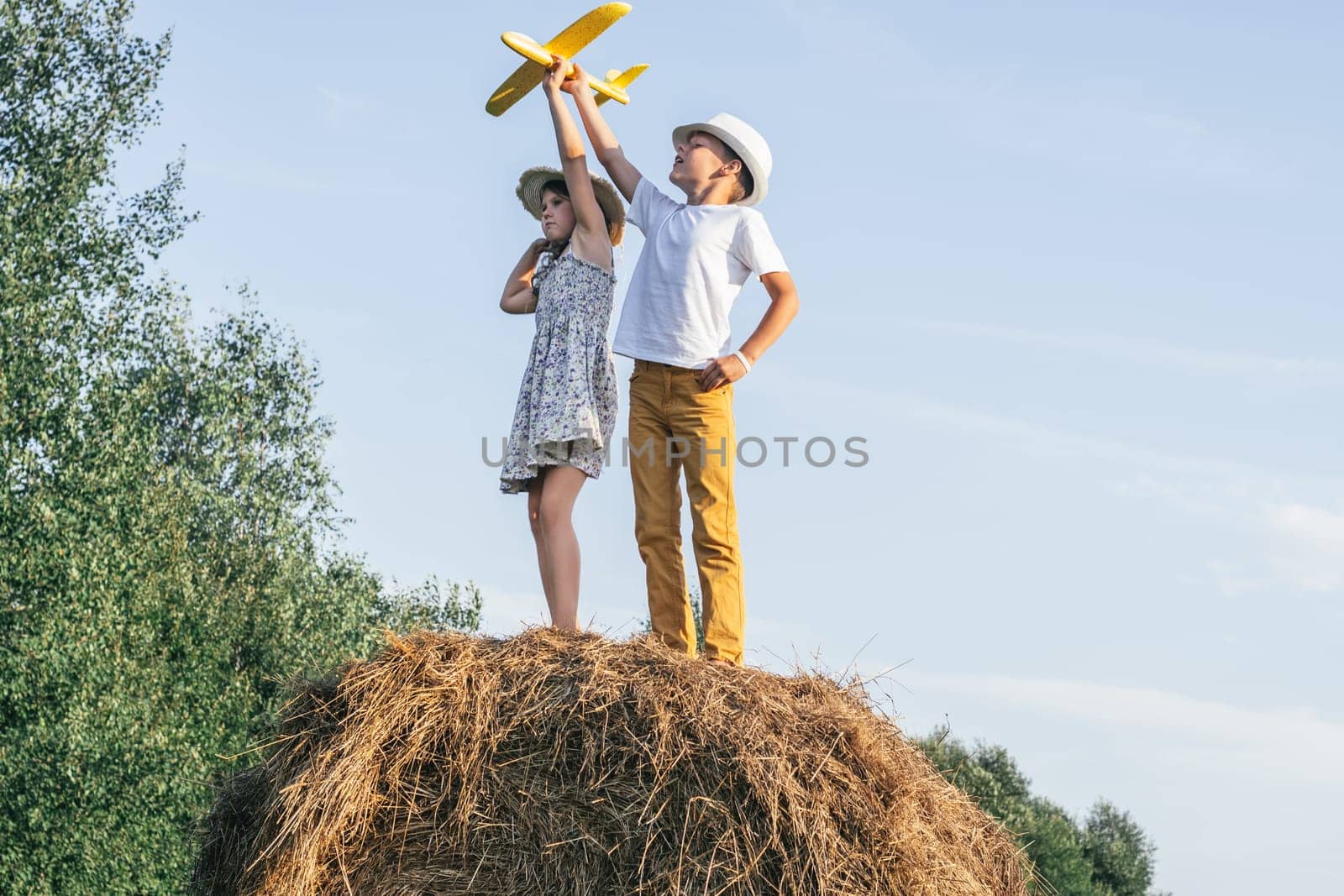 Little girl and boy together launch yellow toy airplane standing at top of haystack. Children on hayrick from below view. Freedom. Beautiful blue sky. Summer vacation, countryside lifestyle.