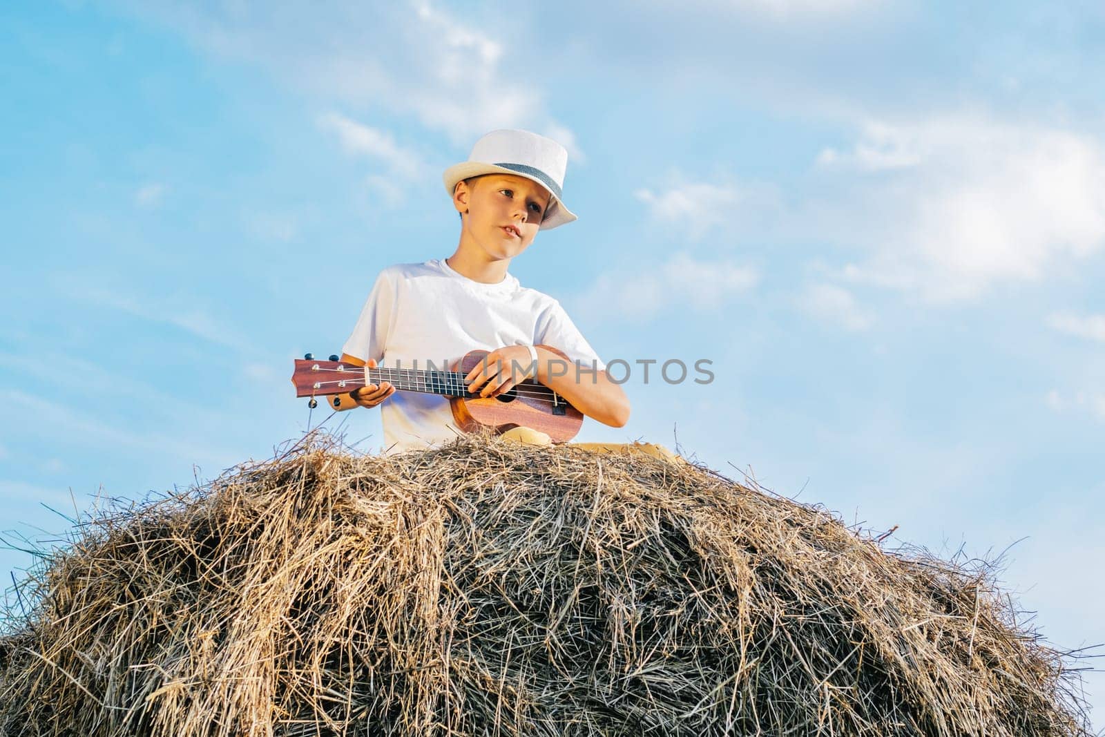 Handsome school aged boy sitting at top of haystack play ukulele. Carefree child resting on hayrick, from below view. Outdoor walking. Beautiful blue sky. Summer vacation, countryside lifestyle.
