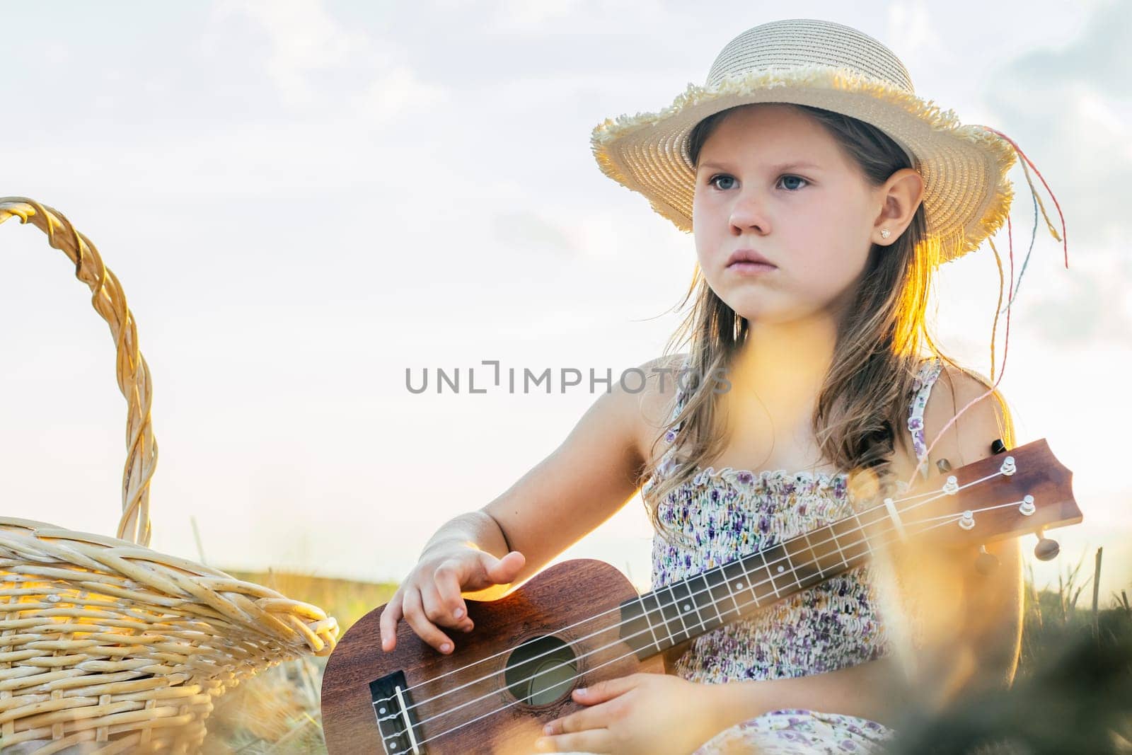 Little girl play ukulele on field. Portrait of child with displeased facial expression in panama. Outdoor picnic. Beautiful sunny sky. Music instrument, hobby. Summer vacation, countryside life.