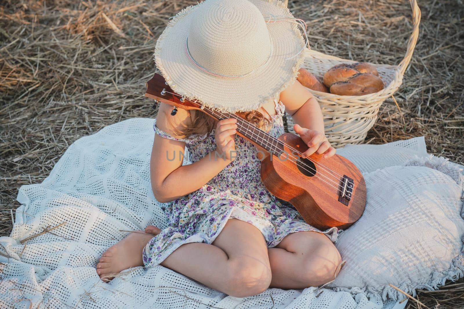 Little girl play ukulele sitting on blanket on field. Portrait of child hiding face under panama hat. Outdoor walking and picnic. Music instrument, hobby. Summer vacation, countryside lifestyle.